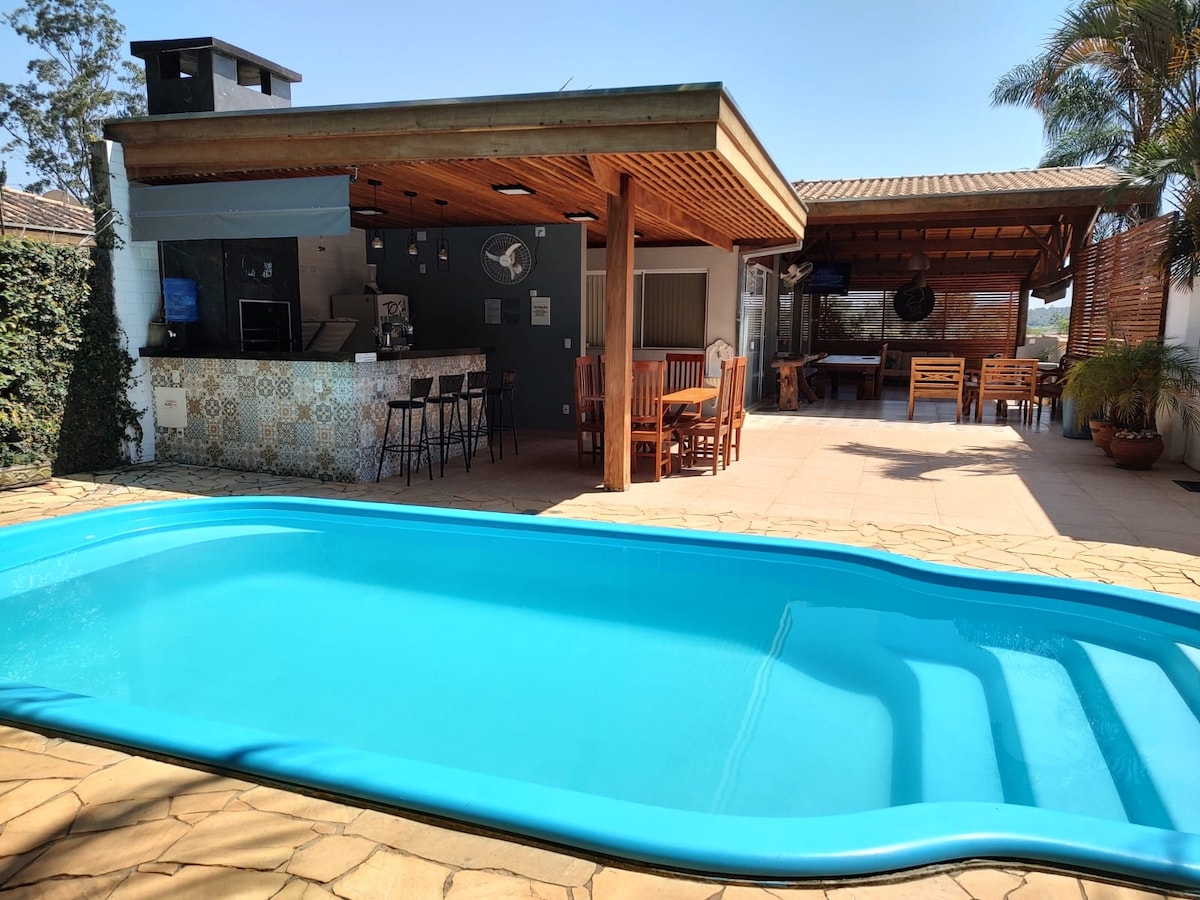 A refreshing swimming pool with gentle steps is positioned in the foreground. A covered outdoor kitchen and dining area are visible, featuring a bar with stools and a spacious wooden table. Lush greenery surrounds the area, enhancing the tranquil ambiance.