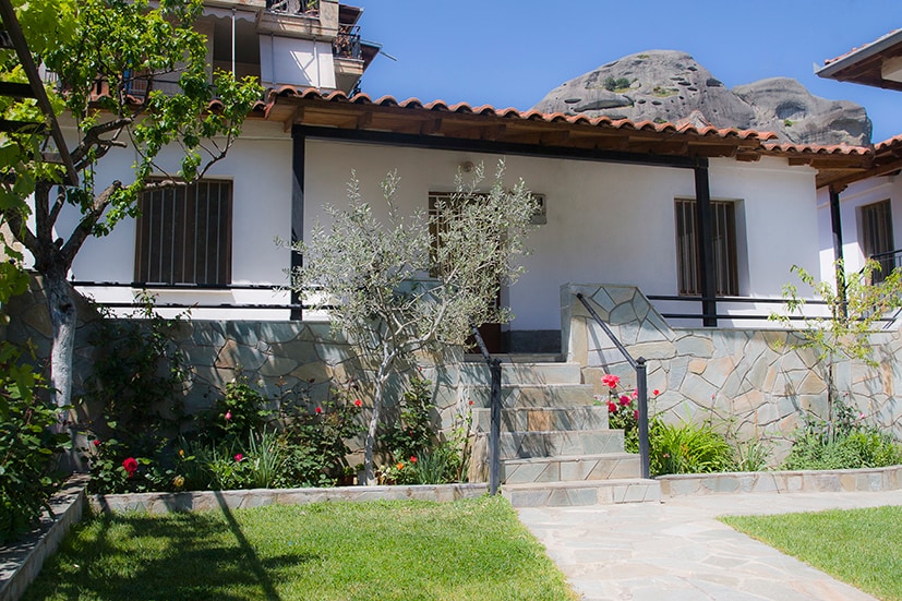 The house features a stone pathway leading up to the entrance, framed by colorful garden plants and flowers. Natural stone accents enhance the exterior, while the terracotta roof complements the clear blue sky above. Windows with simple grilles provide a charming touch.