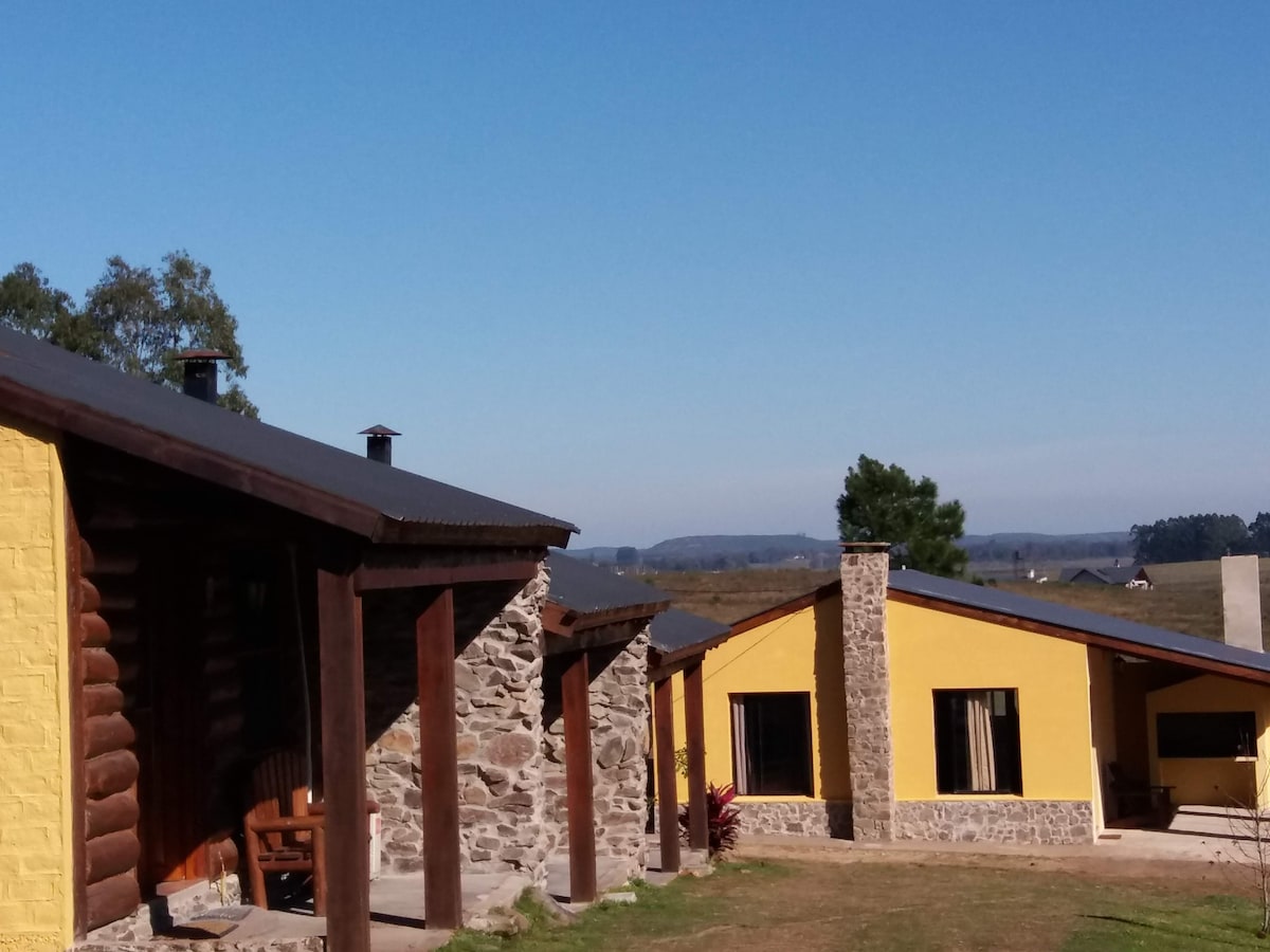 Two cabañas are featured, one with a rustic stone exterior and another with a bright yellow façade. The setting includes a clear blue sky and expansive views of the surrounding landscape, creating a serene country atmosphere. A grassy area connects the structures.