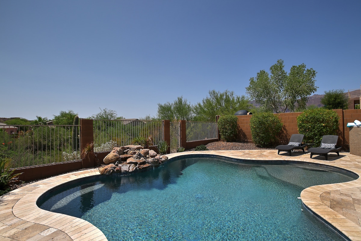 A well-maintained swimming pool glistens under the clear blue sky, framed by a low stone wall and lush greenery. Lounge chairs are positioned nearby, inviting relaxation. Natural rocks create an accent at one end of the pool, enhancing the tranquil ambiance.