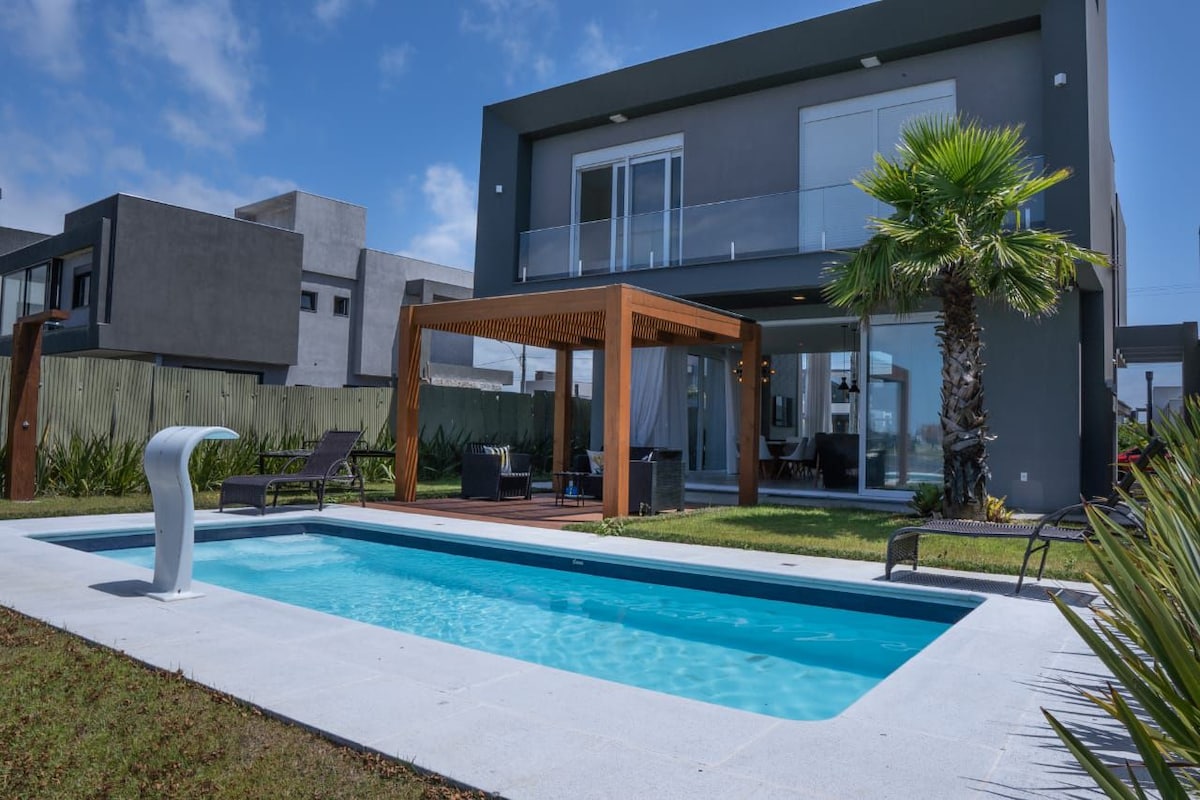 A modern house is set against a clear blue sky, featuring a private pool surrounded by a manicured lawn. A wooden pergola offers shaded seating, while palm trees provide a touch of greenery. Large glass doors reveal a spacious indoor area.