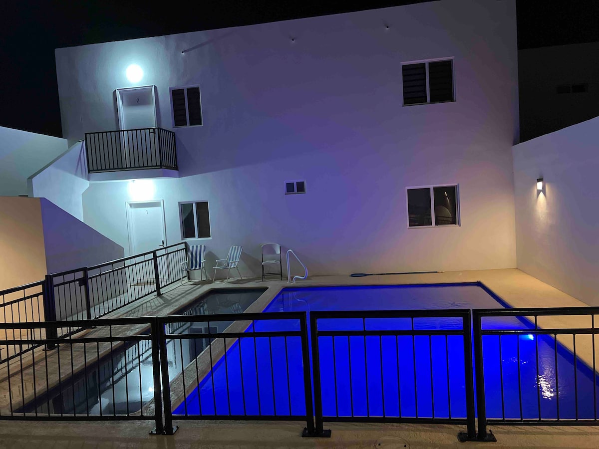 A well-lit outdoor area features two swimming pools, one with a jacuzzi. The surrounding wall is complemented by a safety railing. Three lounge chairs are positioned near the pool area, with a white building visible in the background under soft lighting.