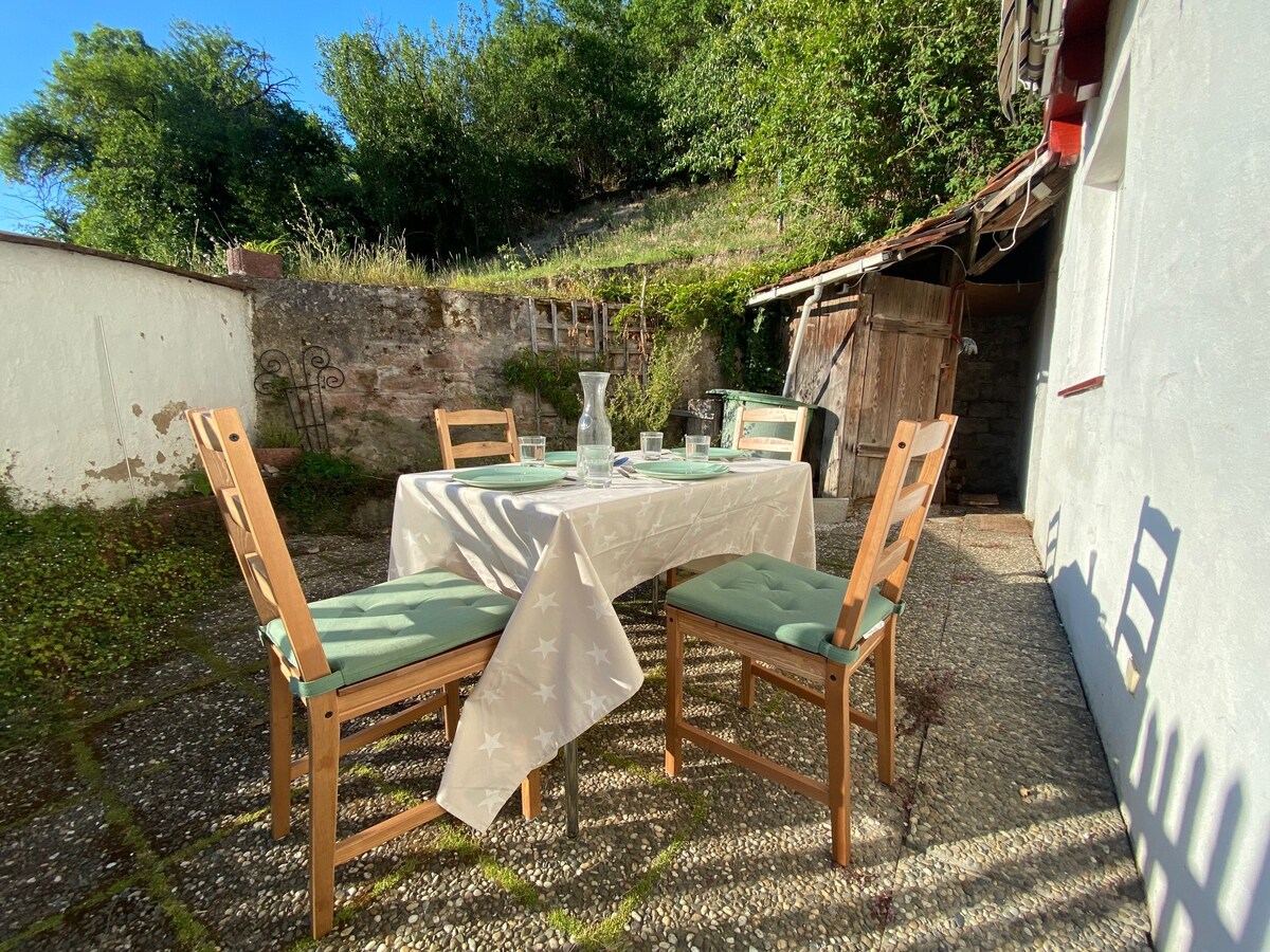 An outdoor dining area features a table set with a neutral-colored tablecloth and glassware. Four wooden chairs with green cushions surround the table. Lush greenery and a stone wall are visible in the background, adding a serene touch to the space.