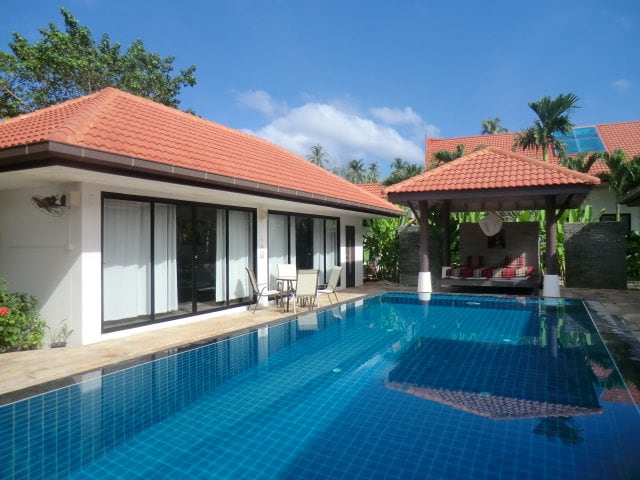The villa features a large, inviting swimming pool surrounded by lush greenery. A shaded sitting area is visible to the right, offering comfortable seating. The exterior of the main building showcases a traditional roof and large sliding doors that open to the pool and garden.
