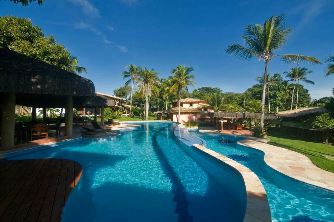 A lagoon-style pool meanders through a tropical landscape, surrounded by lush palm trees. Thatched-roof cabanas and lounge chairs provide shaded areas for relaxation. The bright blue water reflects the clear sky, offering a serene setting for guests to unwind.