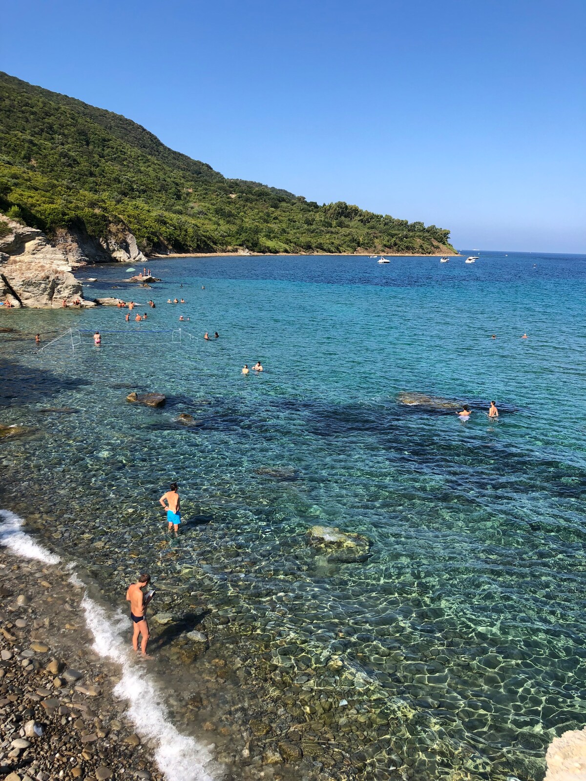 A clear blue sea gently laps onto a pebbled beach. People can be seen swimming and enjoying the water, with rocky cliffs and lush greenery framing the scene. Several boats are anchored in the distance, adding to the serene coastal atmosphere.