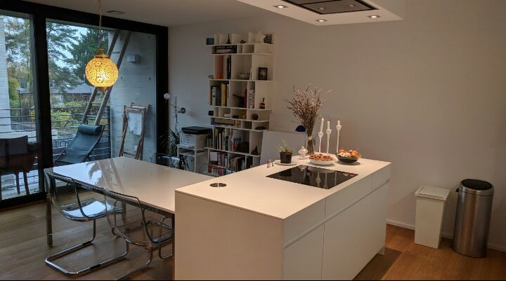 A modern kitchen is showcased, featuring a large white island with an integrated stovetop. A glass dining table is surrounded by chrome chairs. Shelves hold books and decor, while large windows provide natural light and a view of greenery outside.