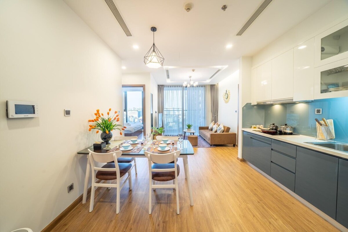 A modern open-concept kitchen and dining area is showcased, featuring a wooden dining table set for four. Plush seating is arranged in the living space, with large windows allowing ample natural light to brighten the interior. Decorative plants add a touch of color to the space.