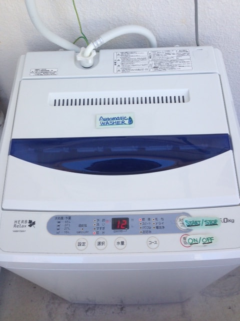 An automatic washing machine is displayed, featuring a top-loading design and control panel. The machine is white with a blue accent on the lid, showing settings for operation. Clear labels for wash functions and a digital display are positioned on the front.