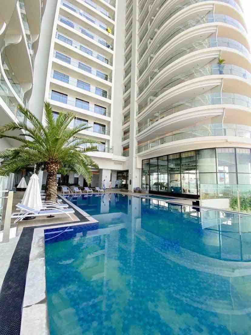 A modern swimming pool area is framed by sleek architectural lines. Several lounge chairs are lined up along the poolside, and palm trees are strategically placed to create a relaxed ambiance. The clear water reflects the building's sleek design and the gentle sunlight overhead.