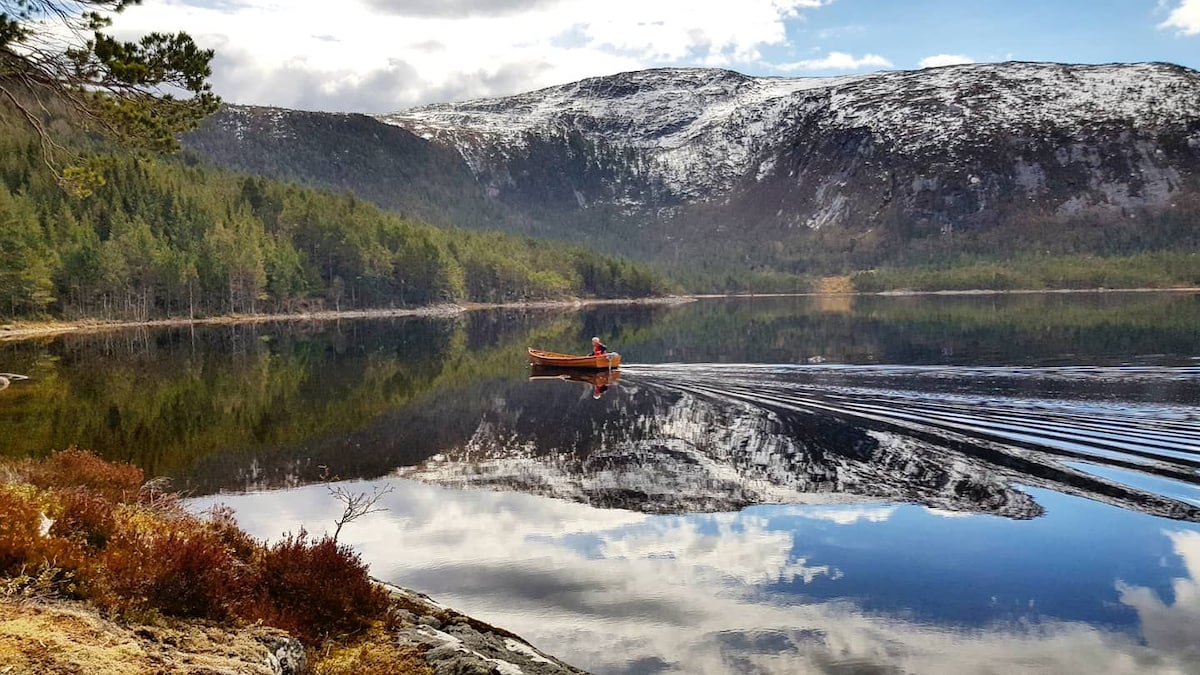 A lone canoe glides across a tranquil lake, reflecting the surrounding mountains and trees. The landscape features a mix of rich greenery and patches of snow, with a clear sky partially covered by clouds. The still waters enhance the serene atmosphere.