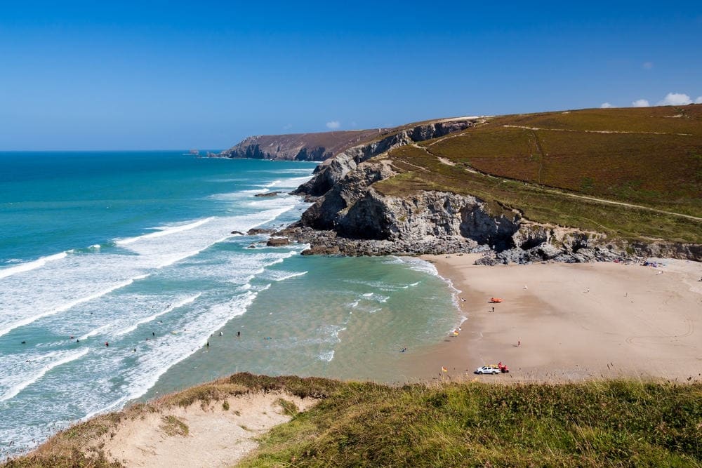 A scenic coastline features sandy beaches gently lapped by the turquoise sea. Rolling hills rise in the background, with a pathway meandering along the cliffs. Sunlight illuminates the water, highlighting surfers and beachgoers, creating an inviting atmosphere for outdoor activities.