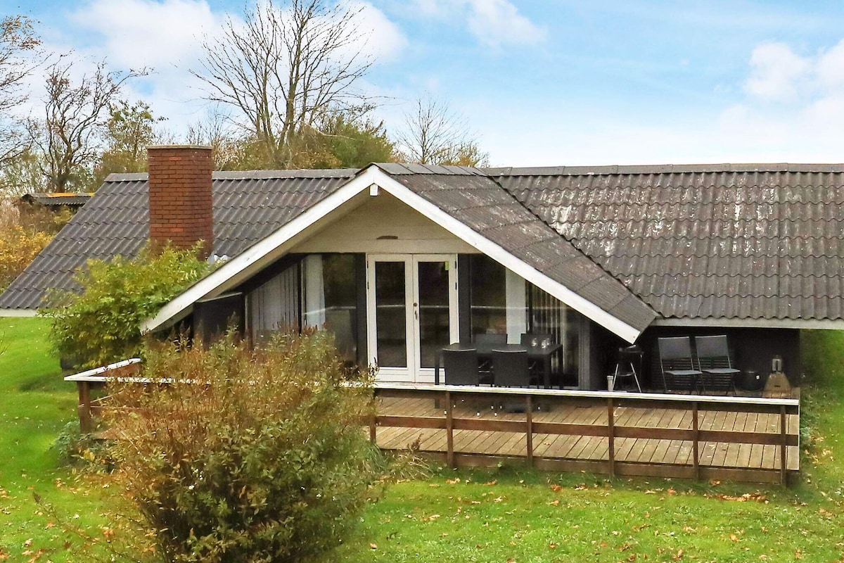 The exterior of a rustic holiday home features a sloping roof and a wooden terrace with seating. Lush greenery surrounds the property, with trees in the background. Large glass doors provide an entrance to the house, creating an inviting connection to the outdoors.