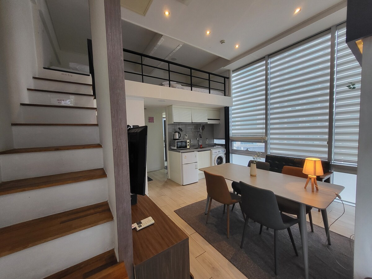 The living area features a light-filled, open layout with large windows covered by horizontal blinds. A dining table accommodates six, complemented by stylish chairs. A staircase leads to the upper level, while a kitchen with modern appliances and a washing machine is visible in the background.