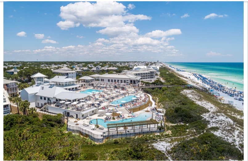 An aerial view captures the WaterColor Beach Club, featuring multiple swimming pools surrounded by lounge chairs. The beach is visible nearby, with white sandy shores meeting gently rolling waves. Lush greenery outlines the area, enhancing the coastal landscape.