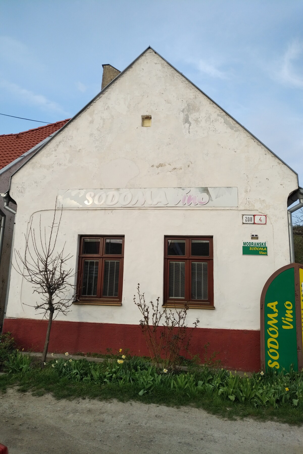 The exterior of a quaint building features a simple gable roof and two large windows with wooden frames. A sign displaying 'Sodoma' is mounted above the windows. Flowering plants adorn the ground, contributing to the outdoor setting.