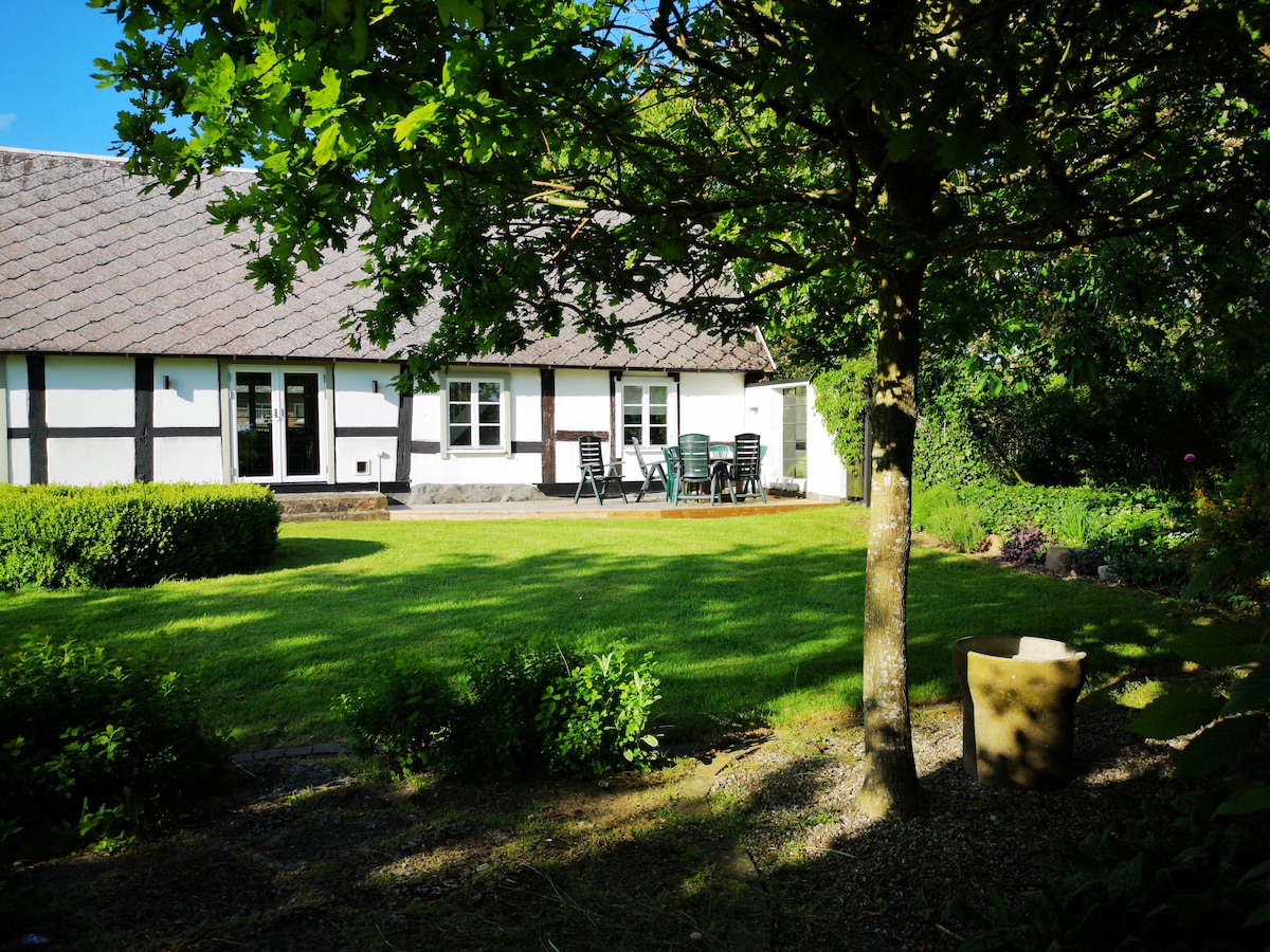 A spacious backyard features a well-maintained lawn surrounded by lush greenery. The exterior of the house showcases a traditional design with visible wooden beams. A patio area with several chairs is seen, inviting outdoor dining and relaxation.