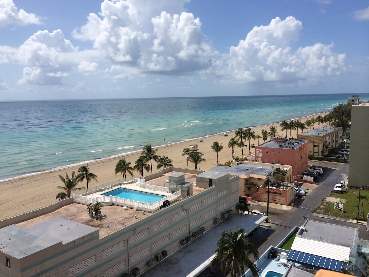 An expansive view of the shoreline displays soft sandy beaches meeting clear turquoise waters. Palm trees gently sway in the breeze, while a nearby pool is visible in the foreground of a low-rise building. The sky is dotted with clouds, adding depth to the scene.