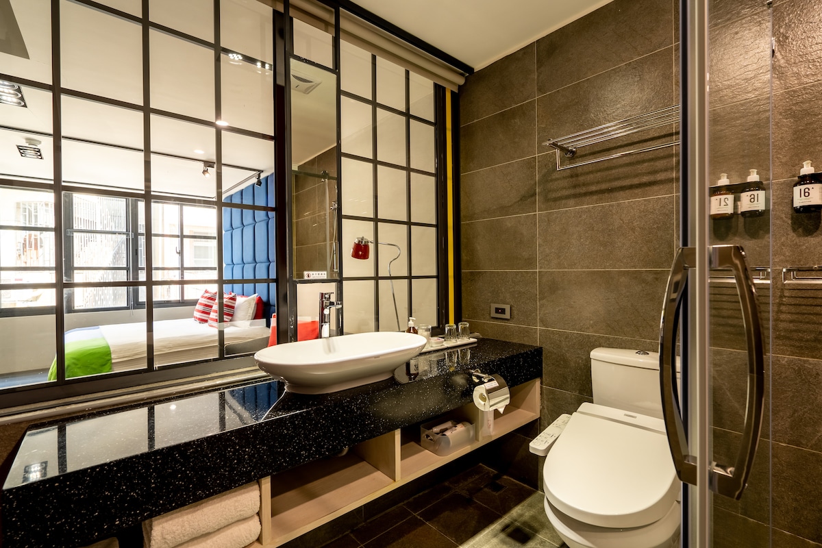 A modern bathroom features a black countertop with a vessel sink and contemporary fixtures. A large mirror with grid patterns reflects the adjacent bedroom. The space includes a white toilet, towel storage below the counter, and wall-mounted racks for toiletries.