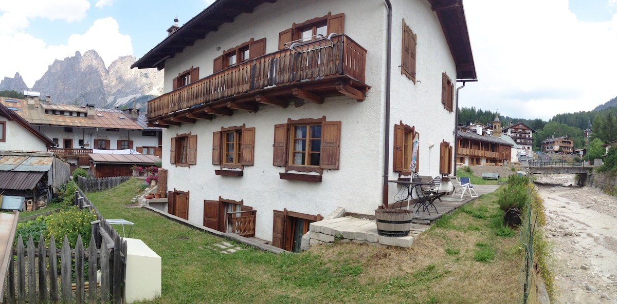 The exterior of the building is depicted, showcasing a traditional mountain home with wooden balconies, framed windows, and white stucco walls. Surrounding greenery is visible, including a garden area and a gently flowing stream nearby. Mountains rise in the background, accentuating the tranquil setting.
