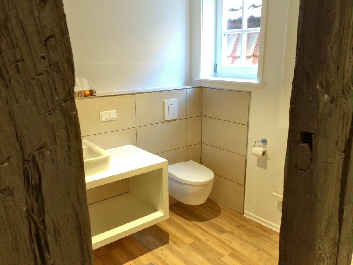 A modern bathroom is shown featuring light-colored tiles and wooden flooring. A wall-mounted sink sits atop a white vanity. A contemporary toilet is positioned beside it. Natural light enters through a small window, enhancing the clean and simple design.