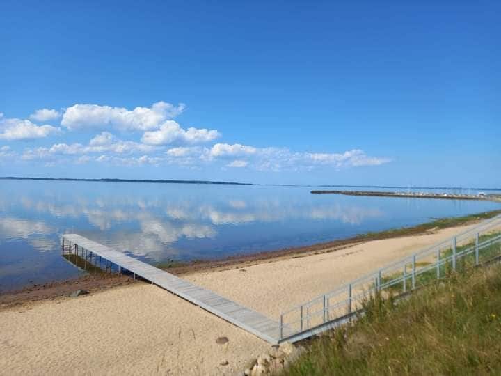A flat wooden pier extends over a calm body of water, reflecting clouds and blue sky. A sandy beach borders the shore, with grassy areas visible in the foreground. The tranquil scene creates a peaceful atmosphere, inviting relaxation and connection with nature.