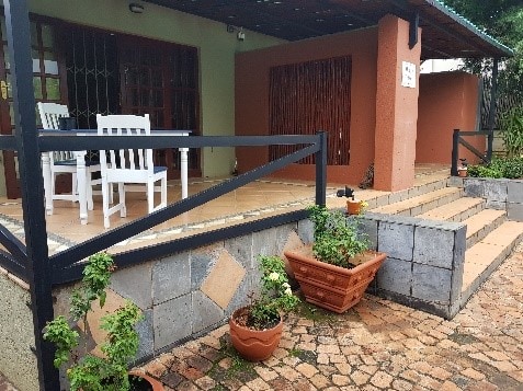 A charming outdoor area is featured, showcasing white wooden chairs arranged on a tiled patio. Potted plants add greenery, and steps lead up to the entrance. The combination of textured surfaces and the natural setting enhances the welcoming aspect of the space.