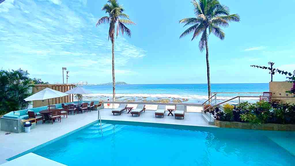 A spacious outdoor area features a clear swimming pool surrounded by lounge chairs, with palm trees providing gentle shade. The beach is visible in the background, highlighting the proximity to the ocean. The setting invites relaxation in a calm environment.