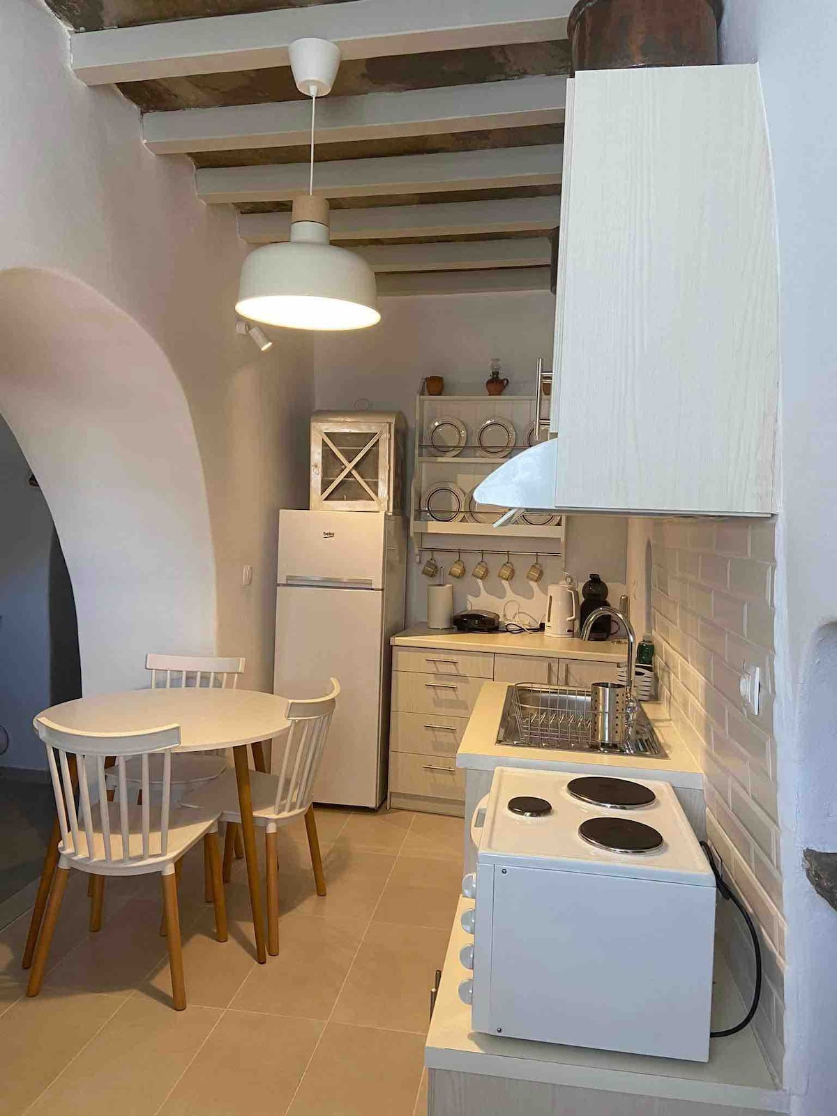 An inviting kitchen area features a round dining table surrounded by four chairs. A refrigerator and an oven are positioned along the wall. Light-colored cabinetry and white tiled walls provide a modern touch, with ample workspace illuminated by a ceiling pendant light.