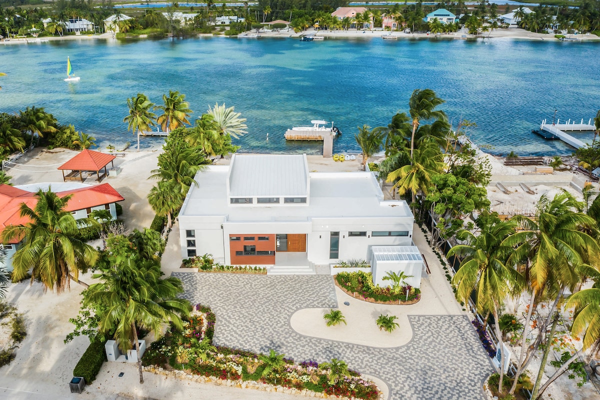 An aerial view of the villa showcases its modern white exterior complemented by vibrant landscaping. Tall palm trees surround the property, which overlooks a tranquil blue cove. Nearby, a private dock and outdoor seating areas can be seen beside the water.