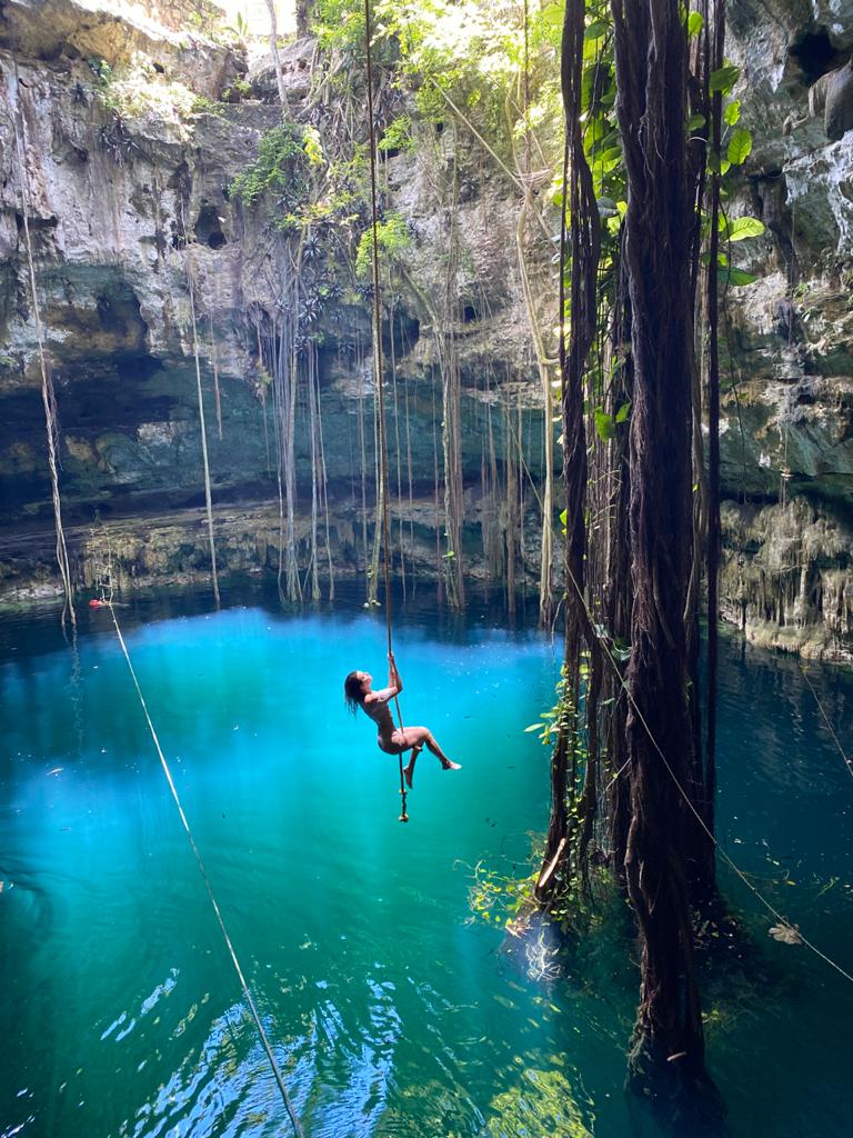 A person swings gracefully from a rope into a cenote surrounded by towering rock walls and lush vegetation. The clear turquoise waters reflect the surrounding greenery, while sunlight filters through the foliage above, creating a serene and enchanting atmosphere.