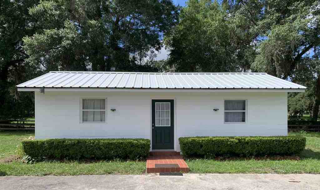A single-story white house is featured, set against a backdrop of lush greenery. A green door is centered on the front, framed by two windows, with a small set of brick steps leading to the entrance. Well-maintained hedges border the foundation.