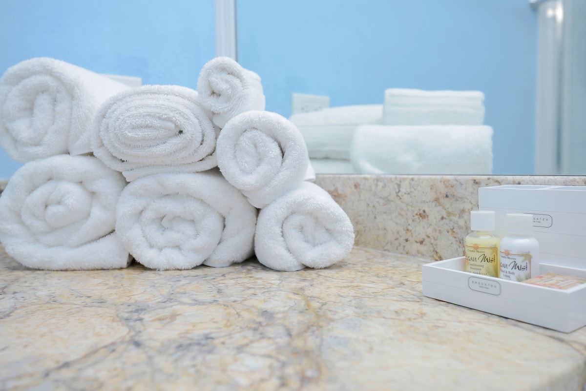 A neatly arranged stack of rolled white towels sits beside a marble countertop. A small selection of complimentary toiletries is displayed in a white box, providing essentials for guests. The background features a light blue wall and a mirror reflecting the serene bathroom environment.