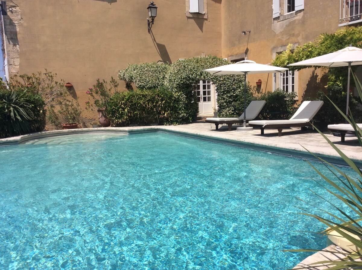 An inviting pool area features clear blue water, surrounded by lounge chairs and sun umbrellas. Lush greenery and flowering plants enhance the space, while the warm exterior walls of the property provide a charming backdrop to the tranquil courtyard setting.