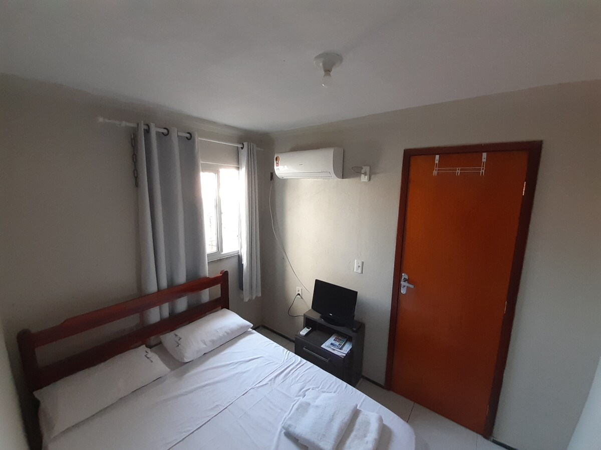 The bedroom features a spacious bed with clean linen and two pillows. A small television is positioned opposite the bed, and an air conditioning unit is mounted on the wall. Natural light enters through a nearby window, with curtains drawn to the side.