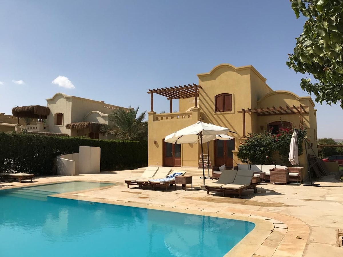 The exterior view of the villa features a private heated pool surrounded by loungers and umbrellas. The building showcases a traditional architectural style with arched windows and a terracotta roof. Lush greenery and palm trees enhance the tranquil outdoor space.