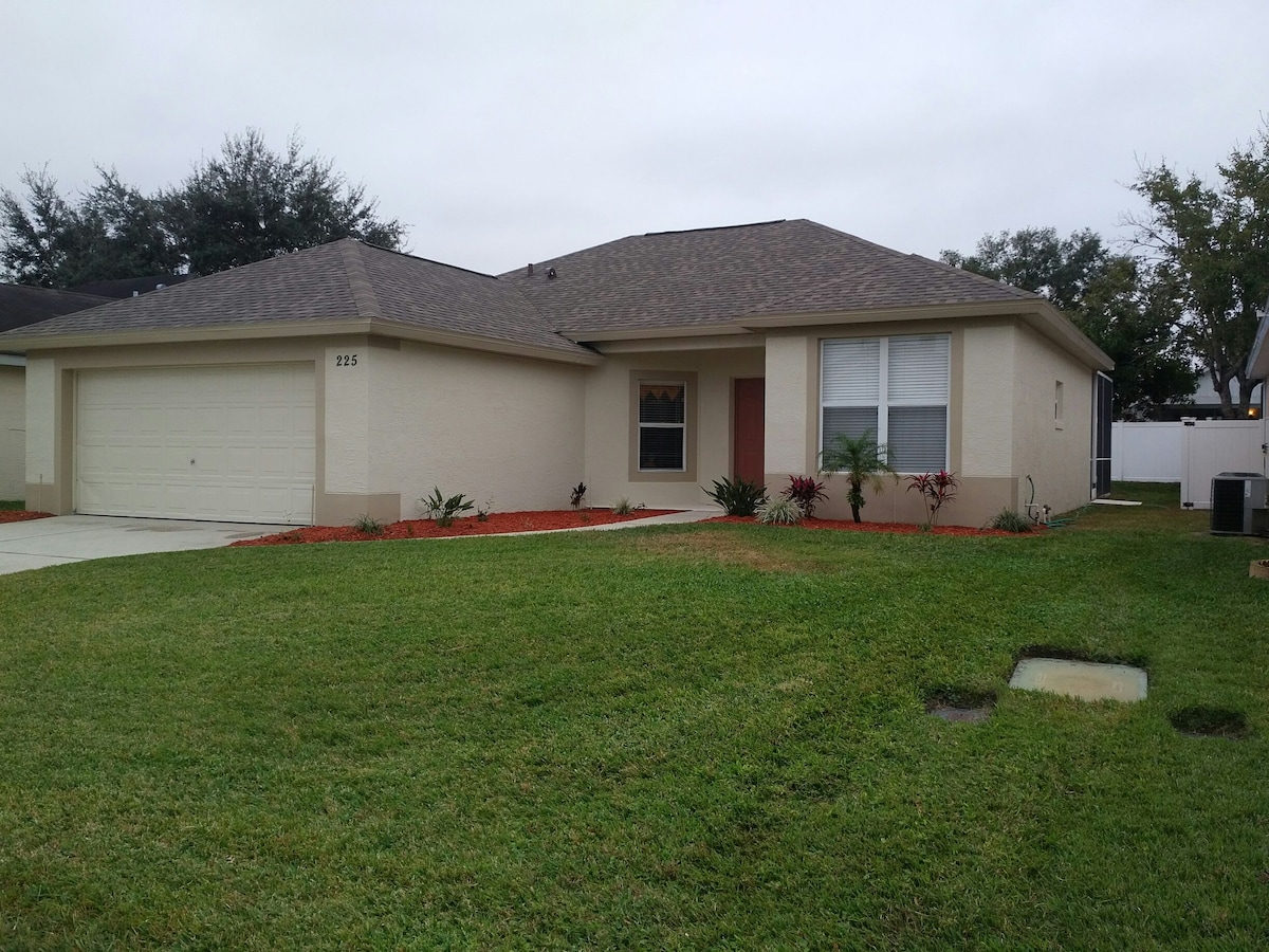 The villa features a single-story exterior with light-colored stucco walls and a patterned shingle roof. A manicured lawn with green grass and landscaped flower beds frames the home, enhancing its curb appeal. A neatly paved driveway leads to the attached garage.