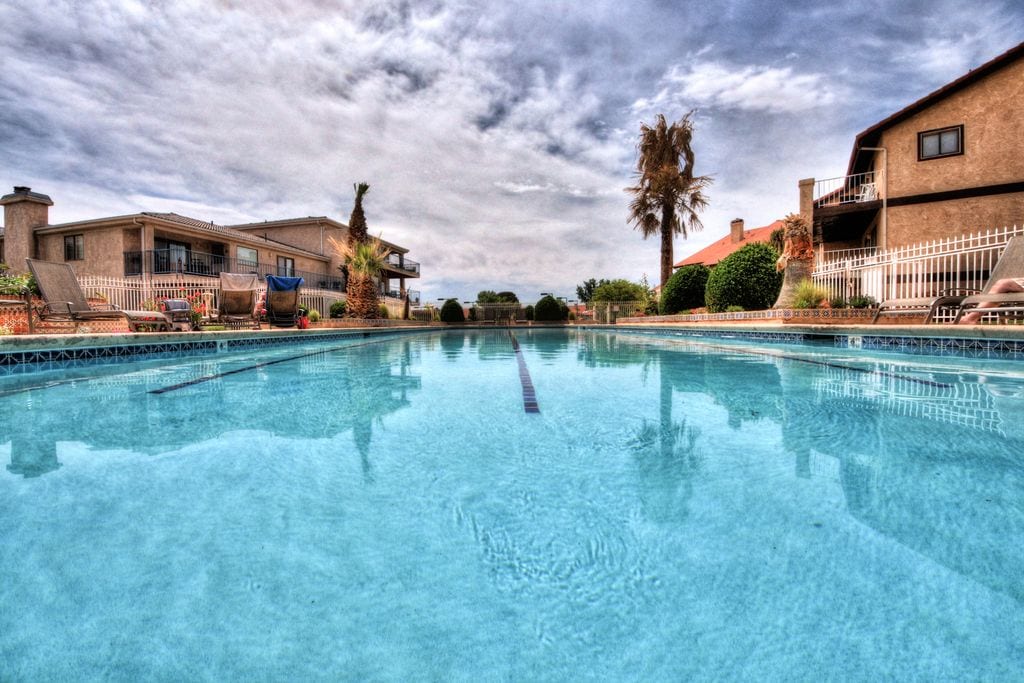 A spacious swimming pool stretches across the image, with clear, inviting water reflecting the sky. Surrounding the pool, lounge chairs are positioned along the deck, while well-maintained landscaping features palm trees and shrubbery, contributing to a relaxed outdoor atmosphere.
