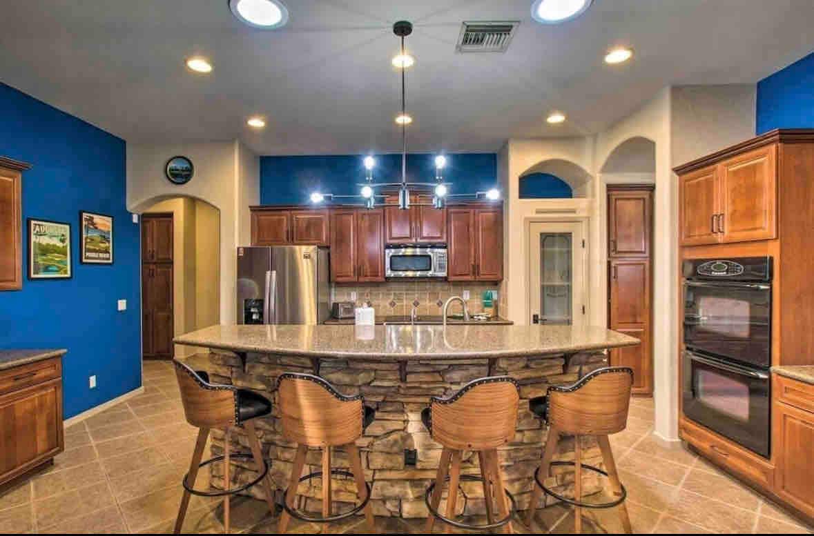 A spacious kitchen features wood cabinetry and a large island with stone accents. Bar stools provide seating along one side. Modern appliances, including a double oven and stainless steel fridge, complement the efficient layout. Soft lighting brightens the room against a bold blue accent wall.