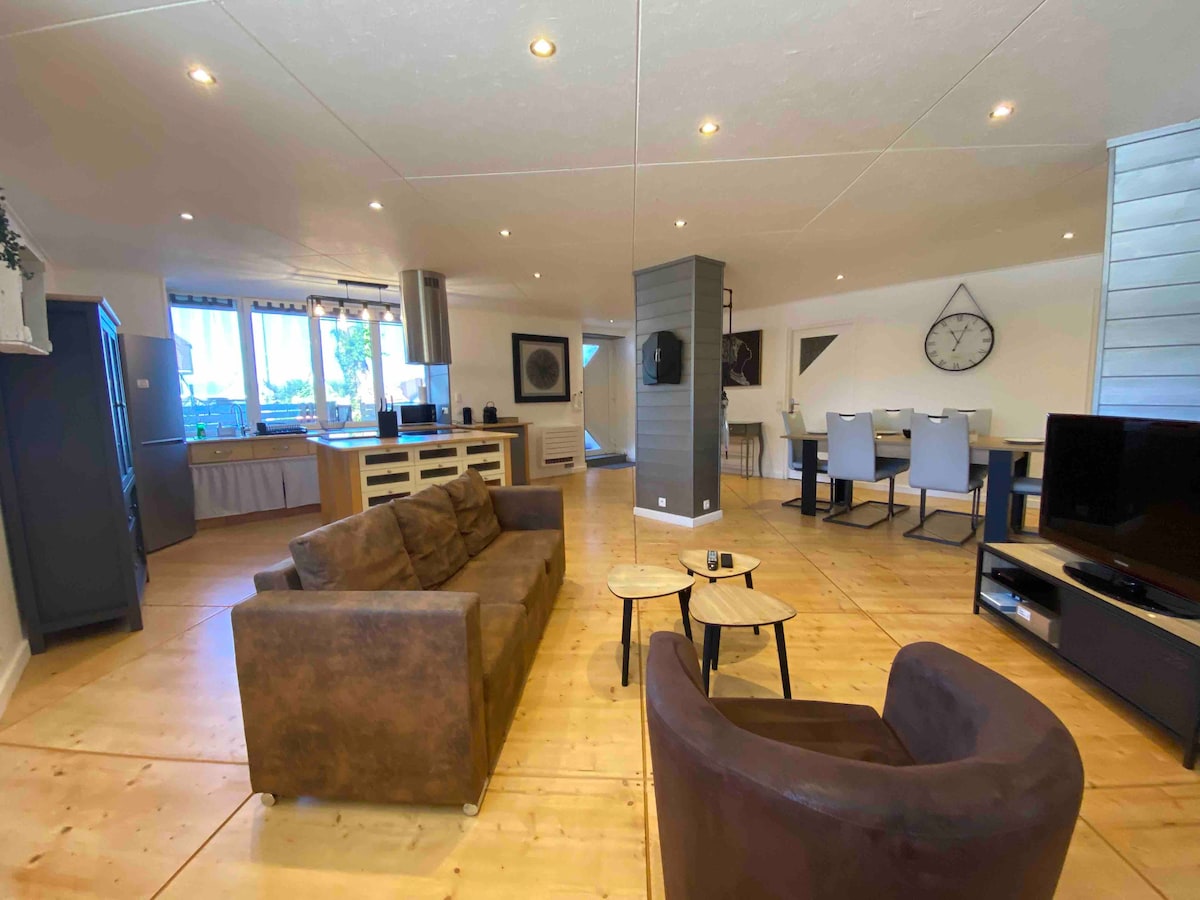 An open-plan living area features a cozy seating arrangement with a brown sofa and an armchair. Light wood flooring complements a modern kitchen area equipped with generous counter space. The dining space is set with a large table surrounded by grey chairs, highlighted by natural light from the windows.