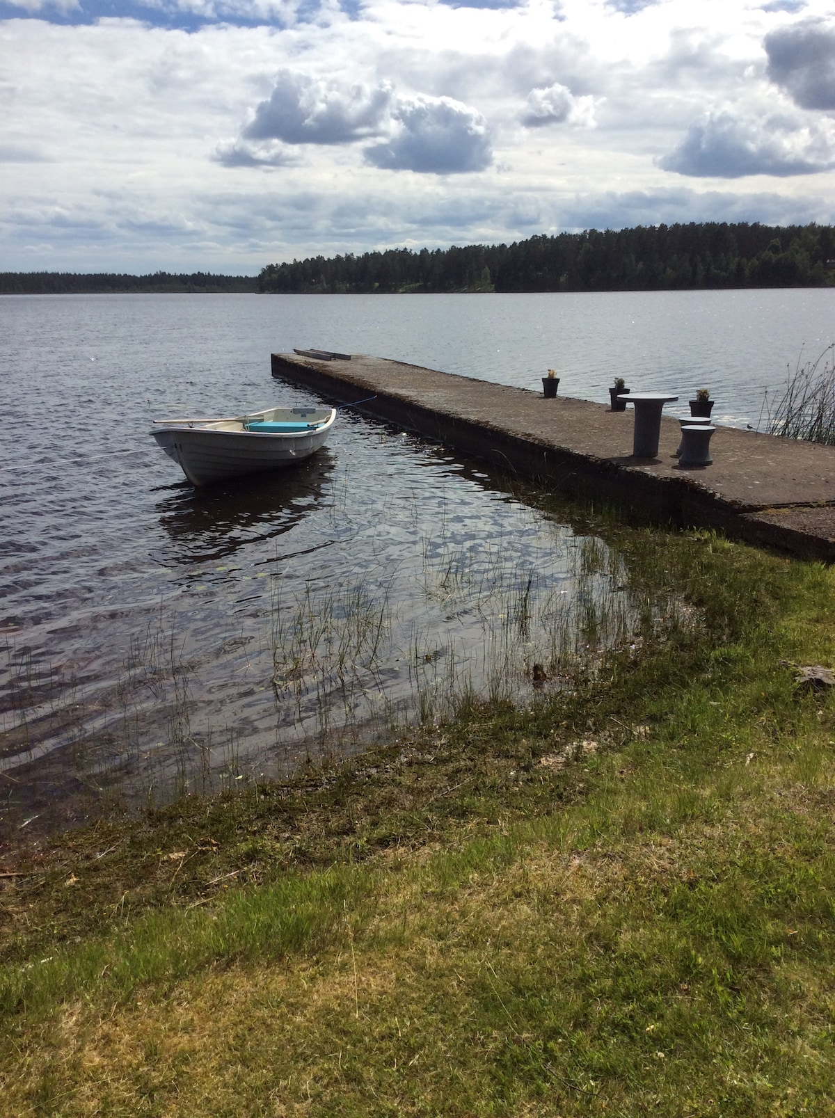 A small boat is anchored next to a concrete pier that extends into the calm water. Lush greenery borders the shoreline, with gentle ripples on the surface reflecting the sky. In the background, trees frame the tranquil landscape beyond the water.