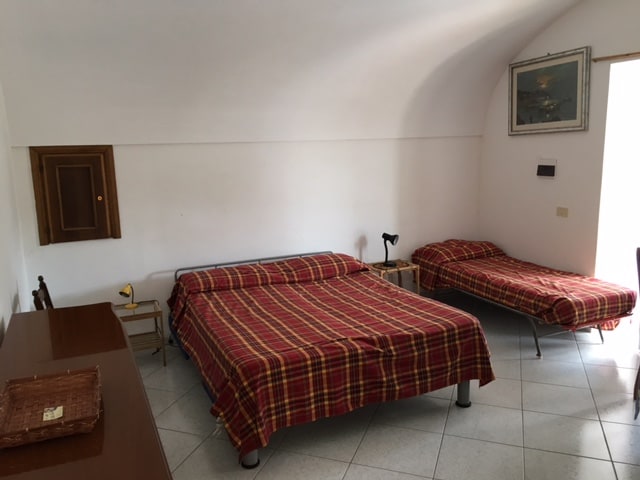 The sleeping area features two spacious beds, one with a plaid covering and the other unmade. A wooden dresser is positioned along one wall, with a lamp beside the larger bed. Soft light fills the room from a window, highlighting the clean tiled floor.