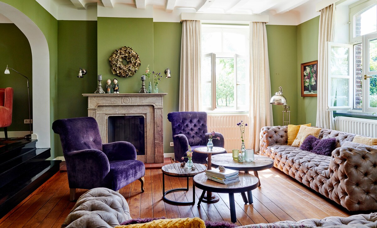 A spacious living area features plush purple armchairs and a comfortable sofa with an array of decorative cushions. Natural light floods through large windows adorned with light drapes, illuminating a charming fireplace and rustic wooden floors.