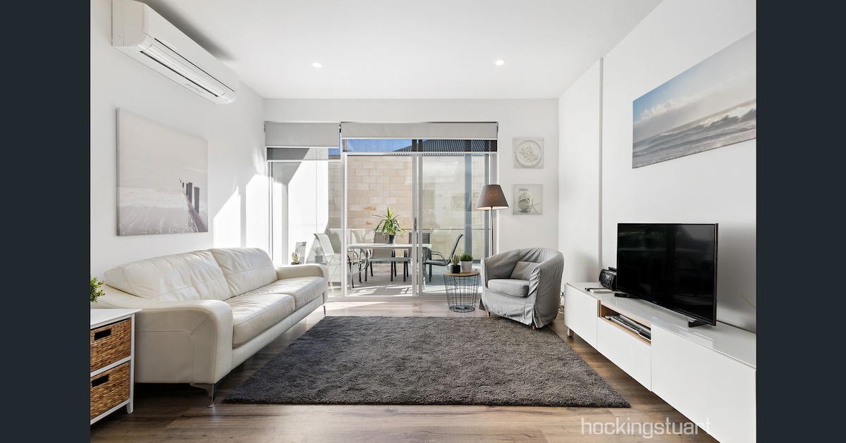A well-lit living area features a spacious white leather couch positioned on a soft gray rug. A comfortable armchair is nearby, and a television is placed on a white media console. Large glass doors allow natural light to illuminate the space, leading to an outdoor seating area.
