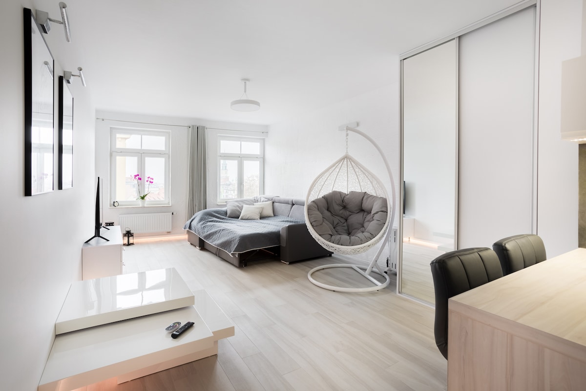A light-filled living space features a spacious bed accompanied by a cozy gray chair suspended from a stand. Large windows offer views of the outdoors, while a small table sits nearby. A modern wardrobe mirrors the bright ambiance of the room.