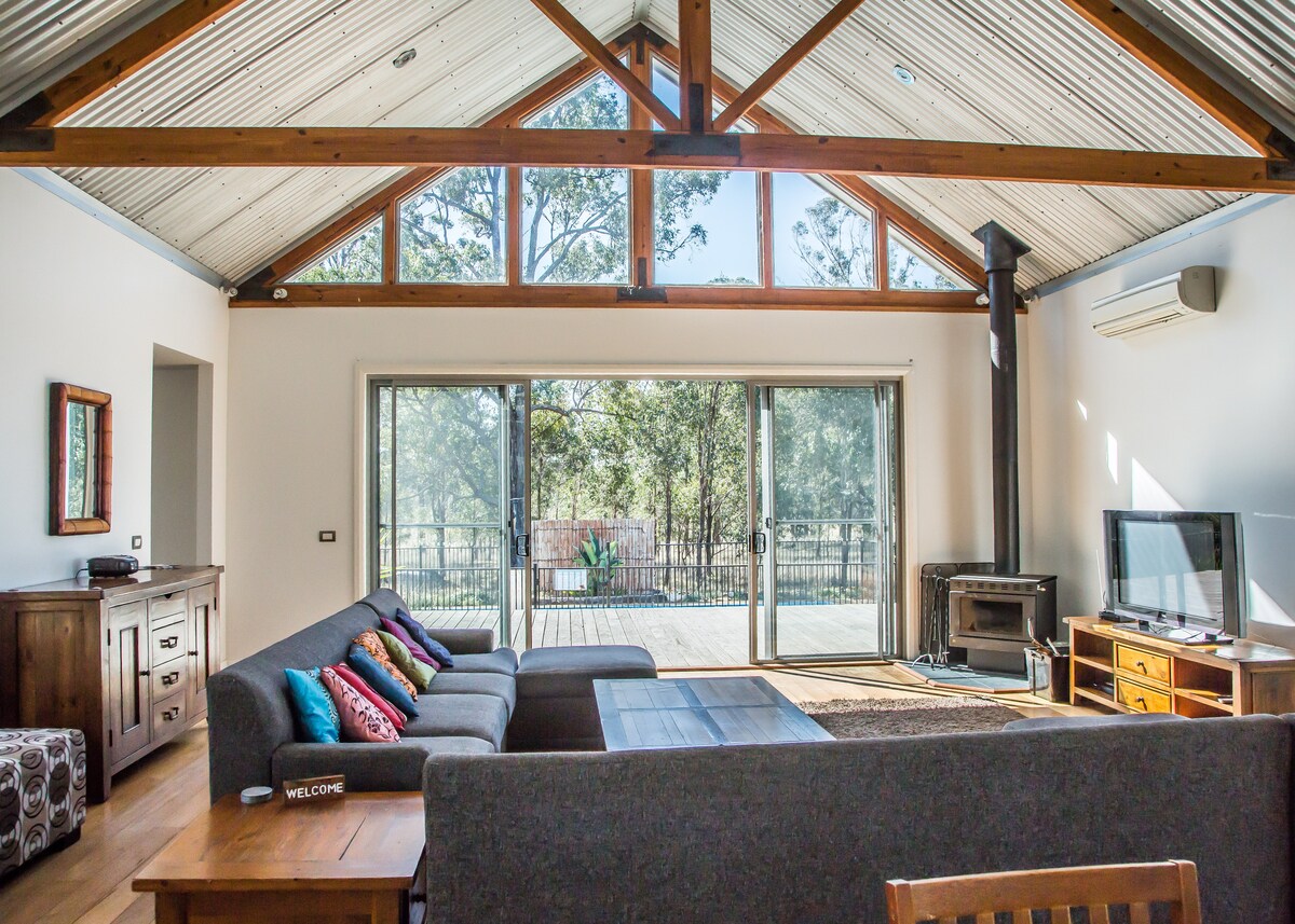 The spacious living area features high vaulted ceilings with timber beams. Large glass doors provide a view of the surrounding nature. A comfortable sectional sofa with colorful cushions faces a wooden coffee table. A television and wood-burning stove are situated against one wall, enhancing the inviting ambiance.