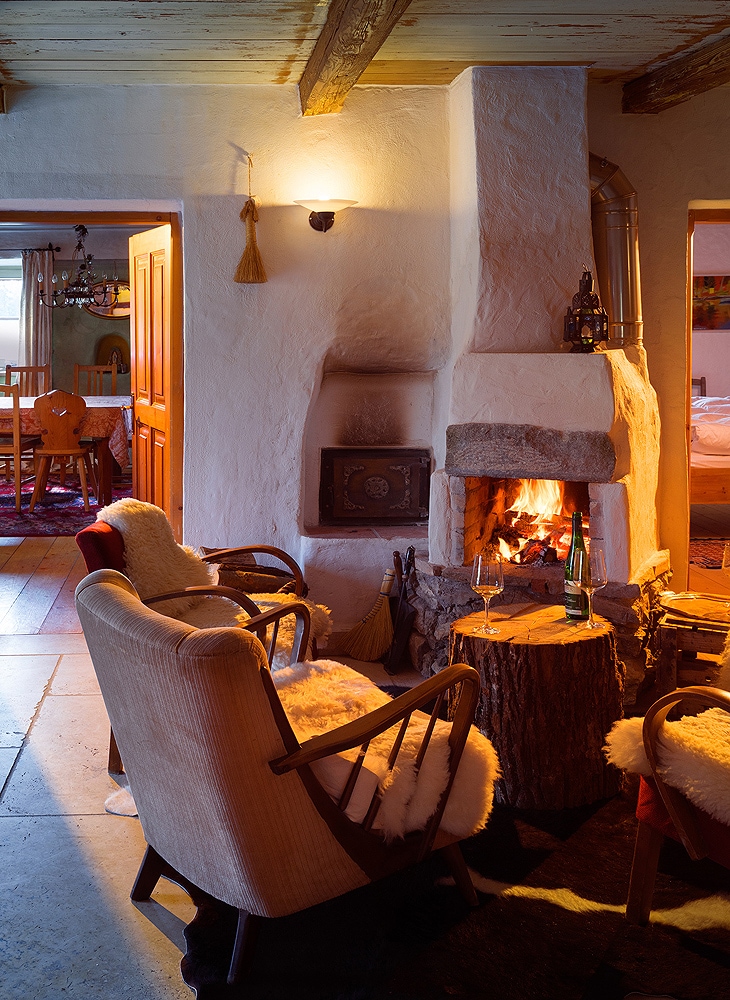 A warm seating area is highlighted by a rustic stone fireplace, showcasing flickering flames. Two cozy armchairs adorned with soft throws face a wooden side table. The space is softly illuminated by ambient lighting, with inviting textures enhancing the overall atmosphere.