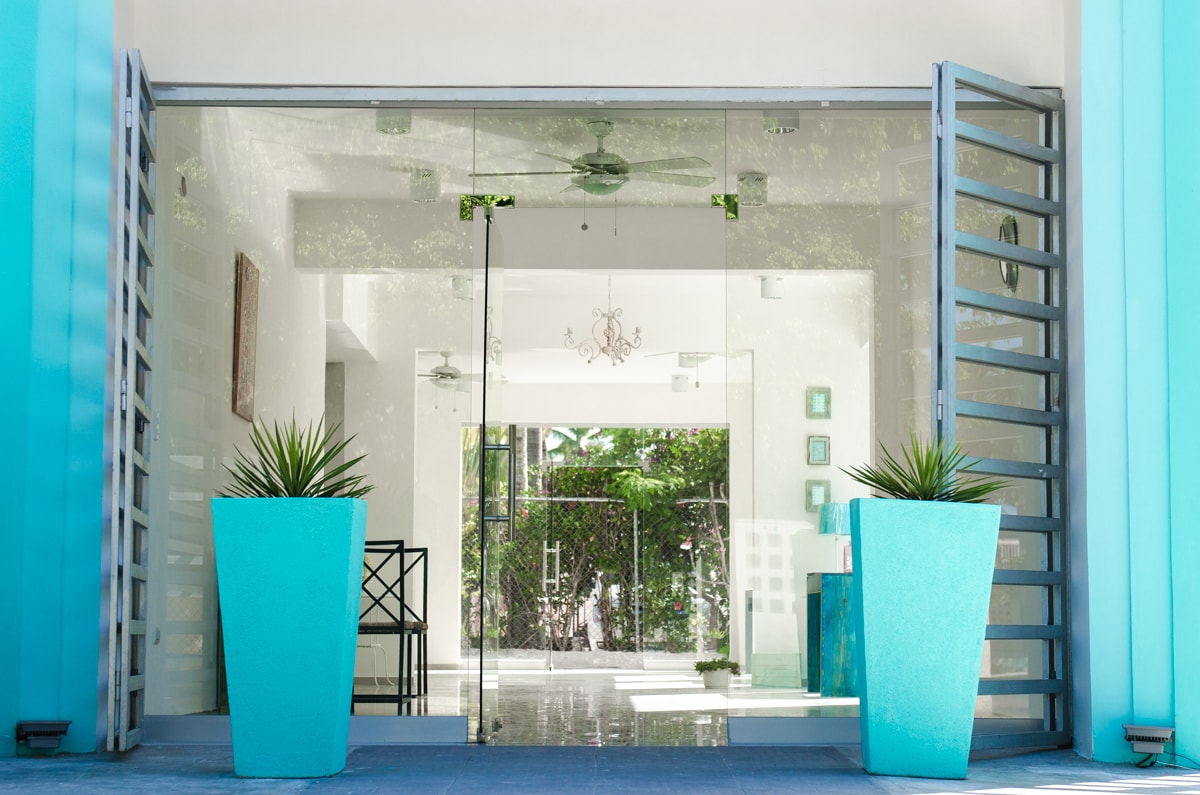 A welcoming entrance features large glass doors flanked by vibrant turquoise planters. The minimalist design is complemented by a polished floor and a glimpse of greenery visible beyond the opening. A black chair is positioned to the side, enhancing the modern aesthetic.