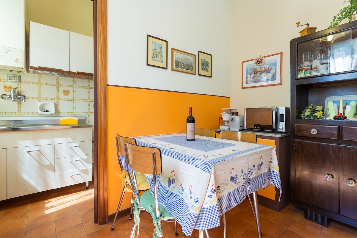 A cozy dining area features a wooden table with a patterned tablecloth and seating for four. A bottle of wine sits in the center. The walls are painted in a warm orange, complemented by framed art. A wooden sideboard and kitchen area are visible in the background.