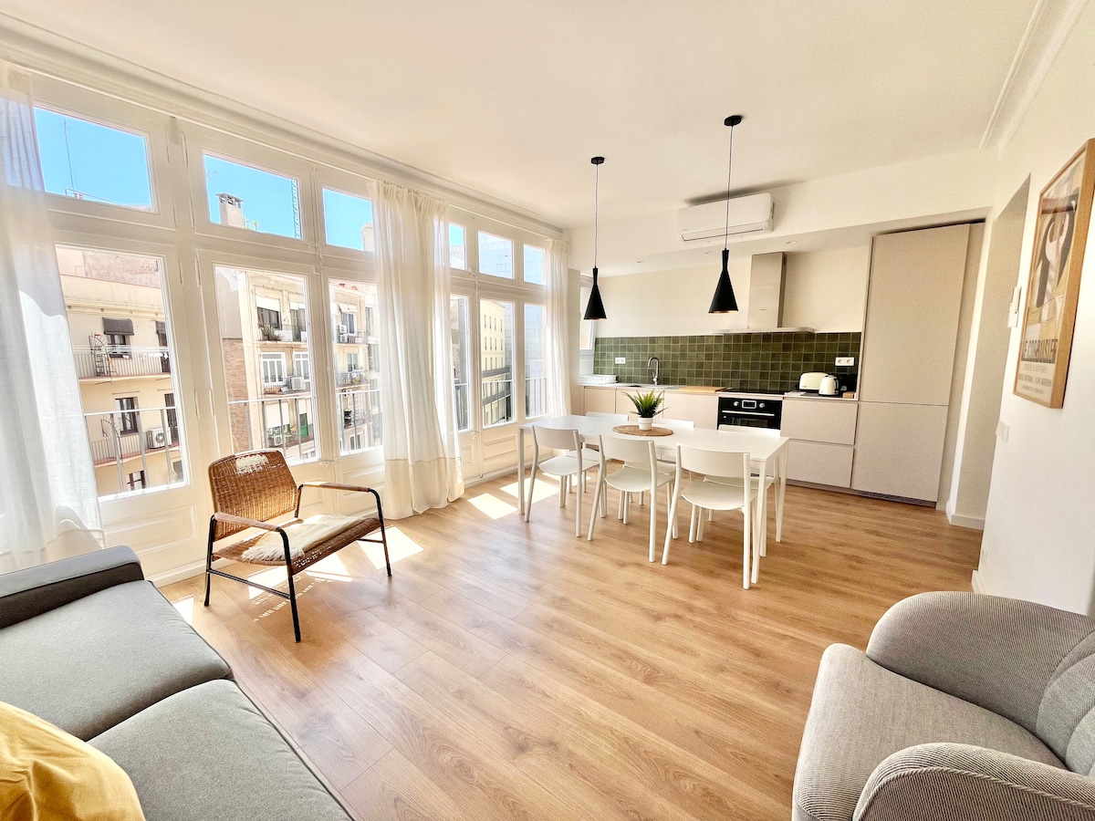 A modern open-plan living area is presented, featuring a light-filled kitchen and dining space. Floor-to-ceiling windows allow natural light to flood in. Simple furniture, including a cozy chair and dining table, enhances the inviting environment, offering a space for relaxation and enjoyment.